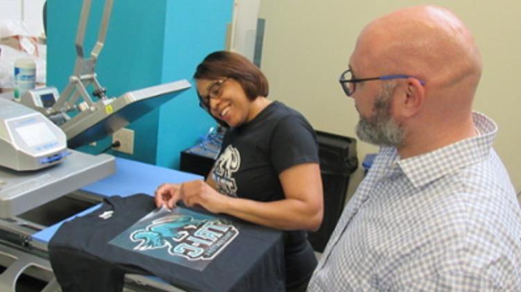 Person in front with his back to the camera in front of a person placing Stomp image on a teal t-shirt